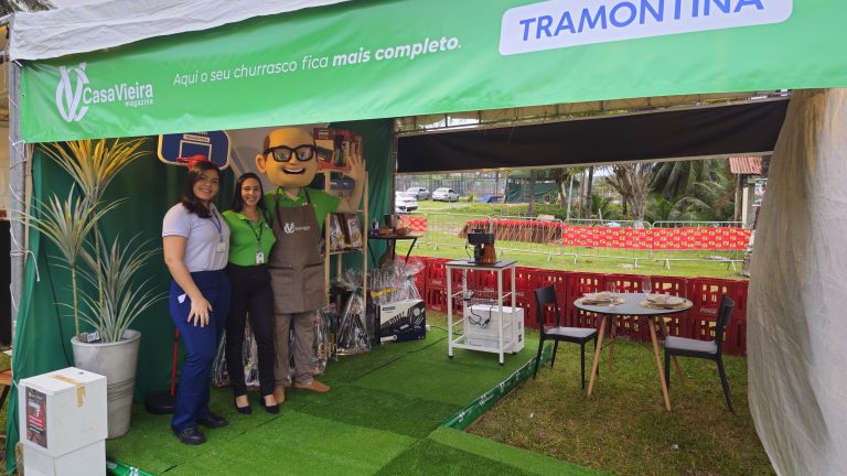 Foto mostrando o stand da Tramontina e da Casa Vieira, com duas funcionárias posando para a foto junto do boneco Vieirão