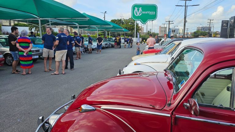 Foto paisagem mostrando parcialmente um fusca vermelho no canto inferior da tela e do lado opostouma fileira de carros antigos expostos com várias pessoas perto