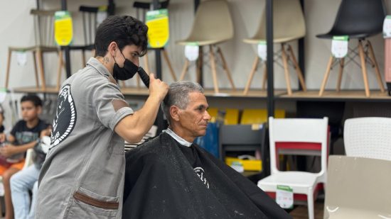 Cabelereiro passando uma máquina de cabelo em um homem adulto, ao fundo é possível perceber que estão dentro de uma loja.